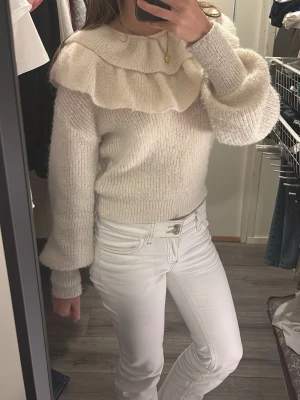 Krämvit stickad tröja med volangkrage - Krämvit stickad tröja med stor volangkrage och långa ärmar. Tröjan har ribbstickade muddar och är gjord i ett tjockt material som känns riktigt mysigt. 