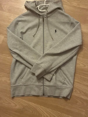 Ralph lauren Tröja - Ljusgrå hoodie med dragkedja från Polo Ralph Lauren. Klassisk design med broderad logga på bröstet, huva med dragsko och två fickor framtill. Mjuk bomullsblandning och ribbade muddar för en skön passform.