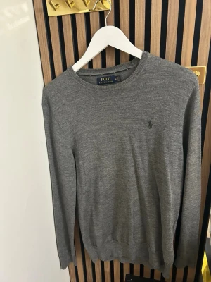 Grå stickad tröja från Polo Ralph Lauren - Snygg grå stickad tröja från Polo Ralph Lauren med rund halsringning och diskret logga broderad på bröstet. Tröjan har långa ärmar och ribbade muddar vid ärmslut och nederkant. Perfekt till lager på lager och enkel att matcha.
