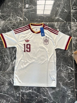 Vit Spanien Adidas fotbollströja - Säljer en vit Spanien fotbollströja från Adidas med vinröda detaljer, nummer 19 på bröstet och klassiska Adidas-ränder på axlarna. Tröjan har gul och vinröd kant vid hals och ärmslut samt broderad landslagssköld. Tillverkad i ett lätt och svalt material.