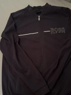 Svart zip hoodie från Hugo Boss - Snygg svart hoodie från Hugo Boss med dragkedja framtill och vit logga på bröstet. Stilsäker design med en vit linje som detalj över bröstet. Tillverkad i mjukt material och har långa ärmar samt ribbade muddar.