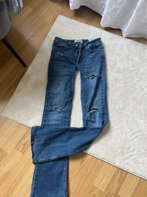 Blå lowboot jeans från ONLY - Säljer ett par blå jeans från ONLY med slitna detaljer och bootcut ben. De är sydda lågmidjat och är därför tightare i låren. Hålen är egengjorda. Jeansen har klassisk femficksdesign och är tillverkade i denim. Perfekta för en avslappnad och trendig look.