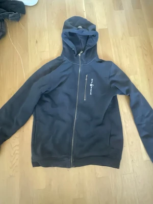 Marinblå hoodie från Sail Racing - Snygg marinblå hoodie från Sail Racing med dragkedja framtill och en bröstficka med logga. Tillverkad i mjukt material med fleecefoder på insidan och huva. Perfekt för chill dagar eller när du vill ha en sportig look.