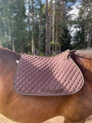 Brunt schabrak med rutmönster - Säljer ett brunt schabrak med quiltat rutmönster och ljusare kantband. Passar perfekt till ridning och ger en snygg look på hästen. Materialet är mjukt och vadderat för extra komfort. Fästen för sadel finns på båda sidor.