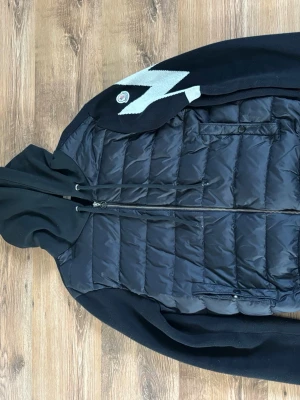 Moncler cardigan  - Snygg svart cardigan från Moncler med quiltad front, huva och dragkedja. Den har ett stort vitt zigzagmönster på axeln och Moncler-logga. Utmärkt för dig som vill ha en sportig och trendig look. Perfekt till hösten som börjar närma sig. Bara skicka om ni har några frågor.