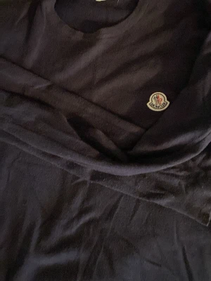Mörkblå stickad tröja från Moncler - Snygg mörkblå stickad tröja från Moncler med rund halsringning och diskret logga på bröstet. Tröjan har långa ärmar och är tillverkad i ett tunt, mjukt material som passar perfekt till lager på lager. Enkel och stilren design. Sitter tight och visar om ni har rn vältränad kropp, lite nopprig men de är den ända defekten   S och xs