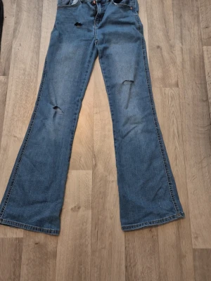 Bootcut jeans med slitningar, blå - Säljer ett par blå bootcut jeans med slitningar på benen och klassisk femficksmodell. Jeansen har en 90-talsinspirerad passform och är tillverkade i denim med synliga sömmar. Perfekta för dig som gillar retrostil och vill ha ett par unika jeans. Det är hål i dom på byxorna på knäna men de e bara att sy därför jag säljer billigt