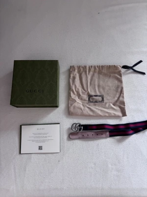 Gucci bälte - Snyggt bälte från Gucci med klassiskt GG-spänne i silverfärg. Bältet har ett randigt band i marinblått och mörklila samt detaljer i ljuslila mocka. Kommer med originalpåse och låda. Perfekt för att lyfta vilken outfit som helst.