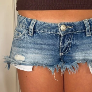 Blå denimshorts med fransar - Säljer ett par blå denimshorts med råa fransar i nederkant och slitna detaljer. Shortsen har klassisk femficksmodell, bälteshällor och knappgylf. Perfekta för varma dagar och ger en avslappnad vibe.