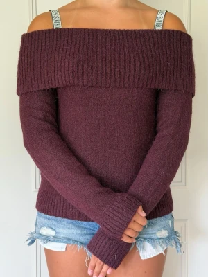 Vinröd off shoulder stickad tröja - Mysig vinröd stickad tröja med bred ribbad off shoulder-krage och långa ärmar. Tröjan har en figurnära passform och ribbade muddar vid ärmslut och nederkant. Perfekt för dig som vill ha en trendig och bekväm look.
