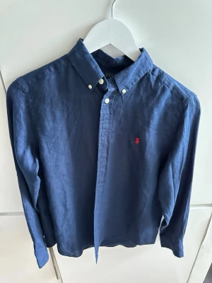 Marinblå linneskjorta från Polo Ralph Lauren - Snygg marinblå skjorta i linne från Polo Ralph Lauren med klassisk button-down krage och röd broderad logga på bröstet. Långärmad modell med normal passform och knappar framtill. Perfekt för en clean och stilren look.