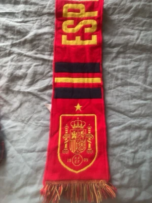 Spain National Football Team Scarf - This is a scarf for the Spain national football team (Selección Española de Fútbol). The scarf features the word 'ESPAÑA' (Spain in Spanish), the team's crest, and the colors of the Spanish flag (red and yellow with some black). The crest includes the initials RFEF (Real Federación Española de Fútbol) and the year 1909, which is the founding year of the Spanish Football Federation. The scarf is likely a fan item used to support the Spanish national team during matches.