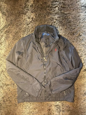 Svart vindjacka från Polo Ralph Lauren - Snygg svart vindjacka från Polo Ralph Lauren med dragkedja framtill och klassisk ståkrage. Jackan har elastiska muddar, två fickor med dragkedja och den ikoniska röda loggan broderad på bröstet. Perfekt för blåsiga dagar och enkel att matcha.