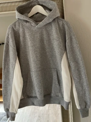 Grå Hollister hoodie med vita detaljer - Snygg grå Hollister hoodie med vita paneler längs sidorna och ärmarna. Klassisk huva och stor magficka framtill. Tillverkad i mjukt sweatshirtmaterial som är perfekt för en avslappnad stil. Ribbade muddar vid ärmslut och nederkant för extra komfort.