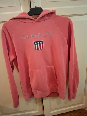 Rosa hoodie från GANT med logga - Snygg rosa hoodie från GANT med broderad logga och amerikanskinspirerat emblem på bröstet. Hoodien har huva, känguruficka och långa ärmar. Perfekt för en avslappnad och sportig stil.