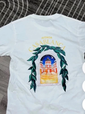 Vit t-shirt med tryck Casablanca - Vit t-shirt med färgglatt tryck fram och stort motiv på ryggen med texten 'Avenida Casablanca' och illustration av en byggnad. Klassisk passform och rund hals. Perfekt för dig som gillar grafiska prints och vill sticka ut lite extra.