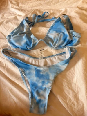 Blåvit bikini från SHEIN - Säljer en snygg blåvit bikini från SHEIN med tie-dye effekt. Överdelen har trekantskupor och smala axelband som knyts i nacken och ryggen. Underdelen är klassisk med låg midja. Perfekt för stranden eller poolen!