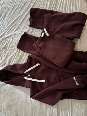 Vinrött set från Russemerch  - Säljer ett vinrött mjukisset från Russemerch med dragkedja och huva. Setet har vita snören och tryckt logga på ärmen. Tillverkat i mjukt bomullsmaterial, perfekt för chill dagar. Både byxor och hoodie ingår. Tröjan har storlek M och byxorna S.