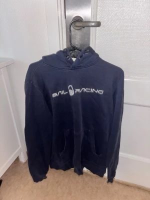 Marinblå hoodie från Sail Racing - Snygg marinblå hoodie från Sail Racing med vit logga tryckt över bröstet. Klassisk känguruficka framtill och justerbar huva med dragsko. Perfekt för dig som gillar en sportig och avslappnad stil. Tillverkad i mjukt bomullsmaterial.