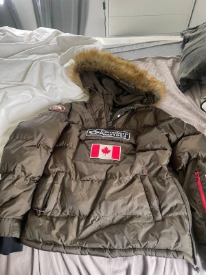 Olivgrön dunjacka Canadian Peak - Olivgrön dunjacka med huva och fuskpäls från Canadian Peak. Jackan har patchar, bland annat en stor kanadensisk flagga och logga på bröstet samt patch på ärmen. Dragkedja och fickor framtill. Perfekt för kalla vinterdagar. 