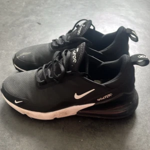 Svarta Nike Air Max 270 sneakers - Säljer ett par svarta Nike Air Max 270 sneakers med vit sula och synlig Air-enhet i hälen. Skorna har en sportig design med vita detaljer och logga på sidan samt texten 'GOLFZIC'. Ovandelen är i textil och syntetmaterial.
