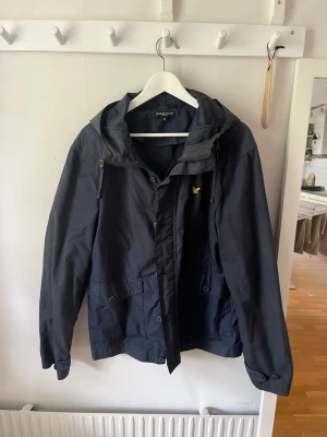 Mörkblå vindjacka med huva - Snygg mörkblå vindjacka från Lyle & Scott med justerbar huva och dragkedja framtill. Jackan har två stora fickor med knappar och en gul logga på bröstet. Perfekt för blåsiga dagar och enkel att matcha till olika outfits.