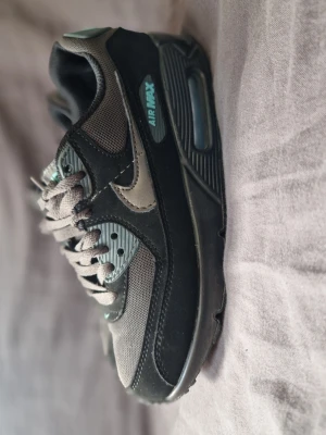 Nike Air Max 90s svarta/grå sneakers - Snygga Nike Air Max 90s sneakers i svart och grått med mesh och läderdetaljer. Skon har den klassiska Air Max-sulan med synlig luftbubbla och turkosa detaljer på sidan och hälen. Perfekt för dig som gillar streetwear och sportig stil.