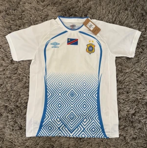 DR Kongo fotbollströja från Umbro - Säljer en vit DR Kongo fotbollströja från Umbro med blå detaljer och geometriskt mönster nedtill. Tröjan har korta ärmar, rund hals och tryckta loggor samt flagga och FECOFA-emblem på bröstet. Perfekt för match eller träning!