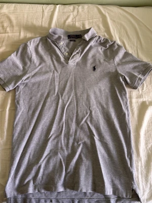 Grå piké från Polo Ralph Lauren - Snygg grå pikétröja från Polo Ralph Lauren med klassisk krage och knappar vid halsen. Tröjan har korta ärmar och den ikoniska loggan broderad på bröstet. Perfekt till jeans eller shorts för en clean och stilren look.