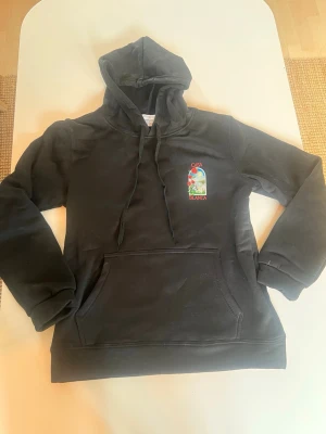 Svart hoodie med Casa Blanca tryck - Säljer en svart hoodie med stor färgglad Casa Blanca-illustration på ryggen och mindre tryck på bröstet. Hoodien har huva med dragsko och en stor magficka. Perfekt för dig som gillar streetwear och unika prints.