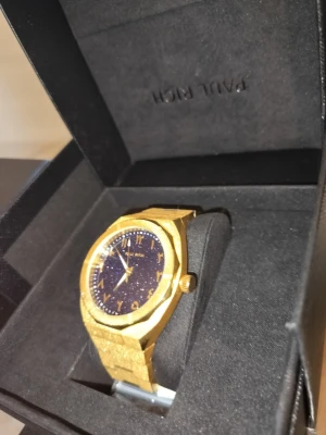 Paul Rich guld klocka med svart urtavla - Snygg klocka från Paul Rich i guld med svart urtavla och guldfärgade detaljer. Klockan har en stilren, kantig form och metallarmband som ger en lyxig känsla. Perfekt accessoar för dig som vill sticka ut och addera lite extra bling till din outfit.