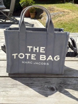 Grå The Tote Bag från Marc Jacobs - Snygg väska i canvas från Marc Jacobs i en stilren grå/blå färg. Väskan har två handtag, axelrem och tydlig vit text framtill med 'THE TOTE BAG'. Perfekt för dig som vill ha en trendig och rymlig väska till vardagen. 💕