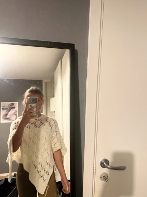 Vit virkad poncho  - Säljer en vit stickad poncho med snyggt blommönster och hålstickad design. Ponchon har en lös och avslappnad passform och är perfekt att slänga över en topp för extra stilpoäng. Materialet känns mjukt och luftigt, vilket gör den skön att bära.