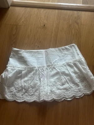 Vit kortkjol med broderade detaljer - Söt vit kortkjol i bomull med broderade blommor och vågig spetskant. Kjolen har en bred midja och vita skjort under