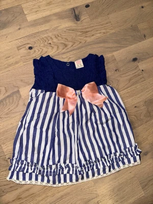 Randig klänning med rosett och volang - Söt kortklänning med marinblå överdel och vit-blåa vertikala ränder på kjolen. Klänningen har en stor rosa rosett framtill och volangdetaljer längst ner samt spetskant. Perfekt för sommarens alla tillfällen.Randig klänning med rosett och volang