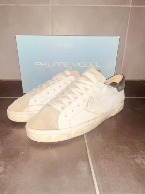 Vita Philippe Model sneakers - Snygga vita sneakers från Philippe Model med beige mockadetaljer och grå häl. Klassisk låg modell med vita skosnören och diskret logga på sidan. Skorna har en stilren design och passar perfekt till streetwear och casual outfits.