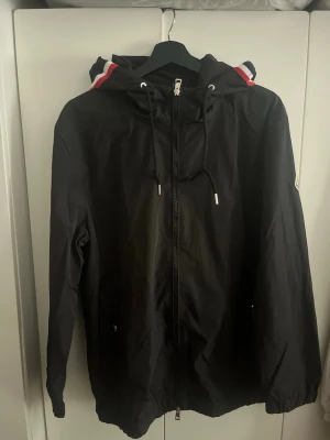 Svart vindjacka från Moncler - Snygg svart vindjacka från Moncler med dragkedja framtill, huva med dragsko och detaljer i rött, vitt och blått. Jackan har två fickor med knappstängning och Moncler-logga på ärmen. Tillverkad i lätt, vattenavvisande material.