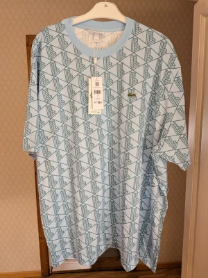 Mönstrad blå t-shirt från Lacoste - Säljer en oversized t-shirt från Lacoste i ljusblått med geometriskt mönster i mörkare blå toner. T-shirten har korta ärmar, rund halsringning och den klassiska Lacoste-loggan broderad på bröstet. Tillverkad i mjuk bomull för skön känsla. Storlek 4XL