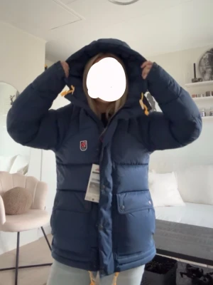 Blå dunjacka med huva från Fjällräven - De bara fråga om fler bilder ❤️( det finns ettt litet hål på höger arm men inget man tänker på eller syns) Snygg blå dunjacka från Fjällräven med stor huva och gula dragskor. Jackan har två stora fickor framtill, diskret Fjällräven-logga på bröstet och dragkedja med vindslå. Perfekt för kalla vinterdagar och riktigt varm.