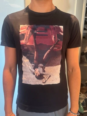 Svart t-shirt med print från Limitato - Svart t-shirt från Limitato med ett stort, färgstarkt print framtill som föreställer en person vid en röd sportbil. Klassisk rund hals och korta ärmar. Tillverkad i mjuk bomull för skön känsla och snygg passform.