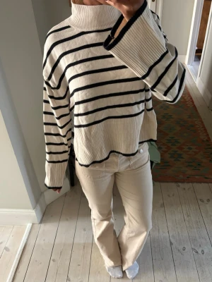 Vit stickad tröja med svarta ränder - Säljer en vit stickad tröja med svarta horisontella ränder. Tröjan har hög krage, vida ärmar och ribbade muddar. Den är oversized och har en avslappnad passform, perfekt för en trendig och chill look. Aldrig använd!
