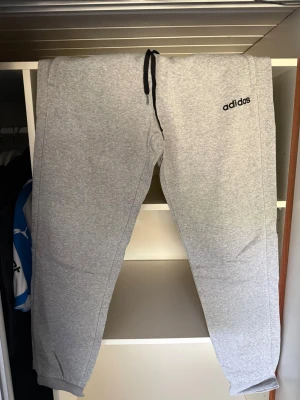 Grå mjukisbyxor från Adidas - Snygga grå mjukisbyxor från Adidas med svart logga på vänster sida. Byxorna har dragsko i midjan och muddar vid bensluten. Tillverkade i mjukt och bekvämt material, perfekta för chill eller träning.