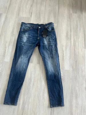 Blå jeans från Dsquared2 - Säljer ett par blå jeans från Dsquared2 ( storlek 54)med cool slitning och färgstänk-detaljer. Jeansen har klassisk femficksmodell, knappgylf och smal passform. Perfekta för dig som gillar en trendig och avslappnad stil. Har använts någon gång men då de it passar mig väljer jag att sälja dem. Etikett finns med.