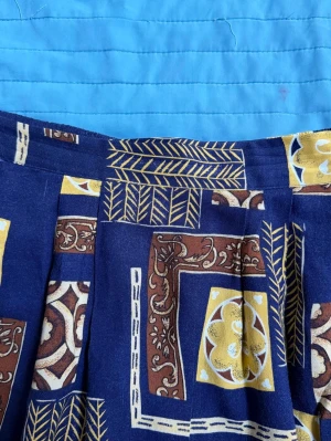 Mönstrad blå vintage  långkjol med gult - Unik vintage långkjol i blått med geometriskt mönster i gult, brunt och vitt. Kjolen har bred linning fram och resår bak för skön passform. Materialet känns som bomull och tyget har ett retroinspirerat tryck med blad och rutor.