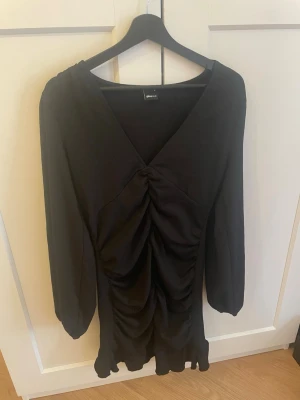 Svart kort klänning med rynk - Snygg svart kort klänning med långa ärmar och v-ringning. Klänningen har rynkade detaljer framtill och volangkant nedtill för en trendig look. Perfekt för dig som gillar stilrena plagg med det lilla extra.