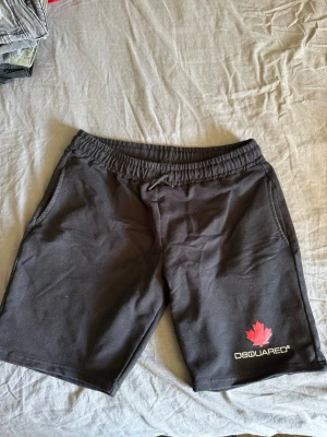 Svarta shorts  Dsquared2 - Säljer ett par svarta shorts Dsquared2 med elastisk midja och snörning. Shortsen har två sidofickor och en röd lönnlöv-detalj samt Dsquared2-logga på vänster ben. Perfekta för sommaren och riktigt sköna att bära.