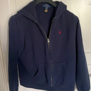 Mörkblå hoodie från Polo Ralph Lauren - Mörkblå hoodie från Polo Ralph Lauren med dragkedja och huva. Klassisk design med röd broderad logga på bröstet och två fickor framtill. Perfekt för när du vill ha en stilren och bekväm look. Passar dig som har storlek S. Skriv till mig om du har frågor