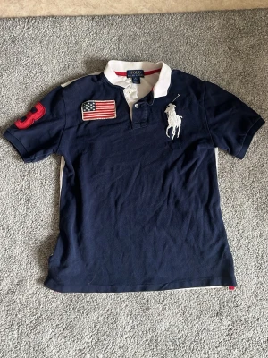 Marinblå pikétröja från Polo Ralph Lauren - Marinblå pikétröja från Polo Ralph Lauren med vit krage och vita detaljer på ryggen. Tröjan har amerikansk flagga och stor logga på bröstet samt röd siffra på ärmen. Klassisk piké med korta ärmar och knappar vid halsen.