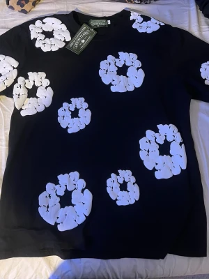Svart t-shirt med vita blommor Denim Tears - Säljer en svart t-shirt från Denim Tears med stora vita blommönster tryckta över hela tröjan. Klassisk rund hals och korta ärmar. Perfekt statement-plagg för dig som gillar streetwear och unika prints.