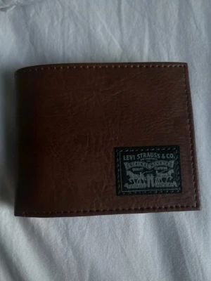 plånbok Levi's  - Snygg brun plånbok från Levi's i äkta läder med klassisk logga på framsidan. Plånboken har flera kortfack och ett större fack för sedlar. Perfekt för dig som vill ha en stilren och praktisk accessoar.